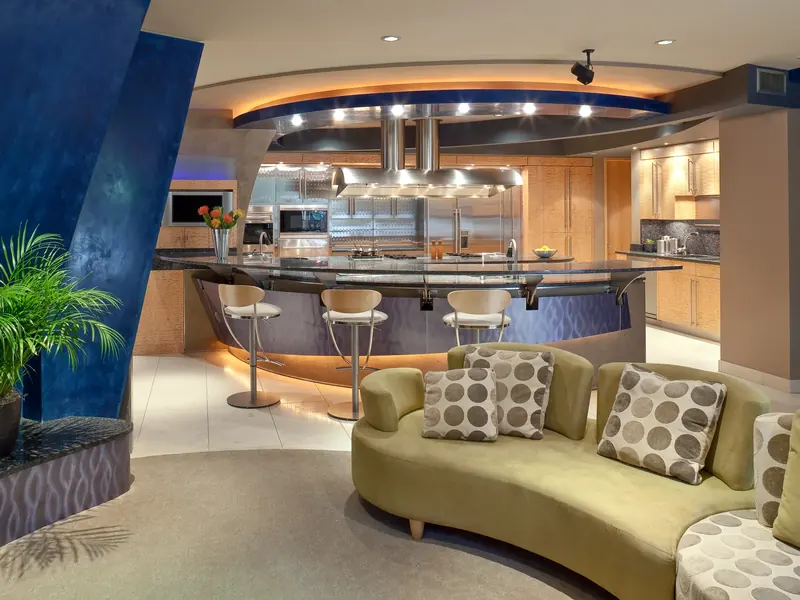 Kitchen beyond a blue plaster column and green sofa: curved bar with stools, stainless hood and a sculpted ceiling ring
