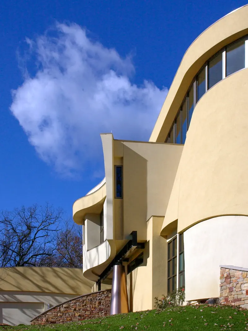 Low view of curved stucco volumes, a cantilevered bay and a tapered metal column at the entry, with a cloud overhead