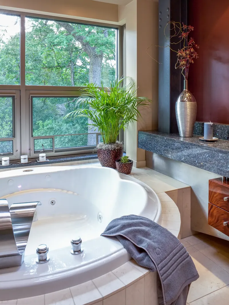 Whirlpool tub beside a window into green treetops, with a potted palm and silver vase