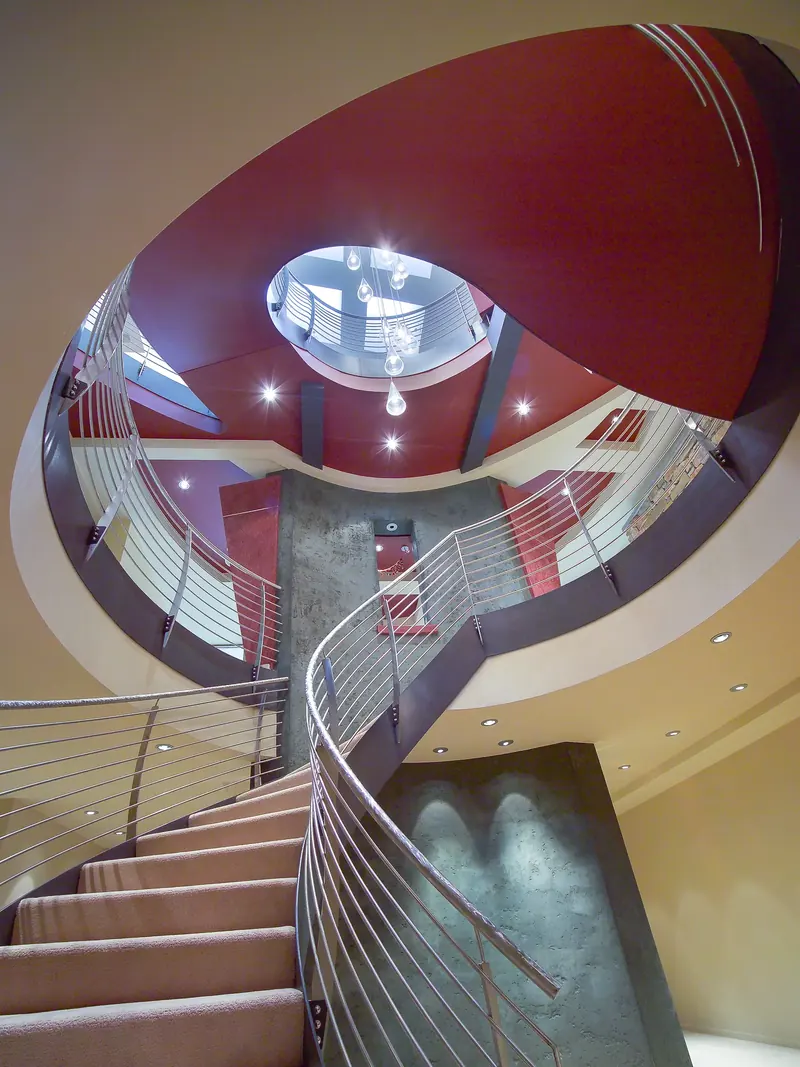 Lower stair curving up into an oval red opening, grey plaster wall and hanging glass lights above