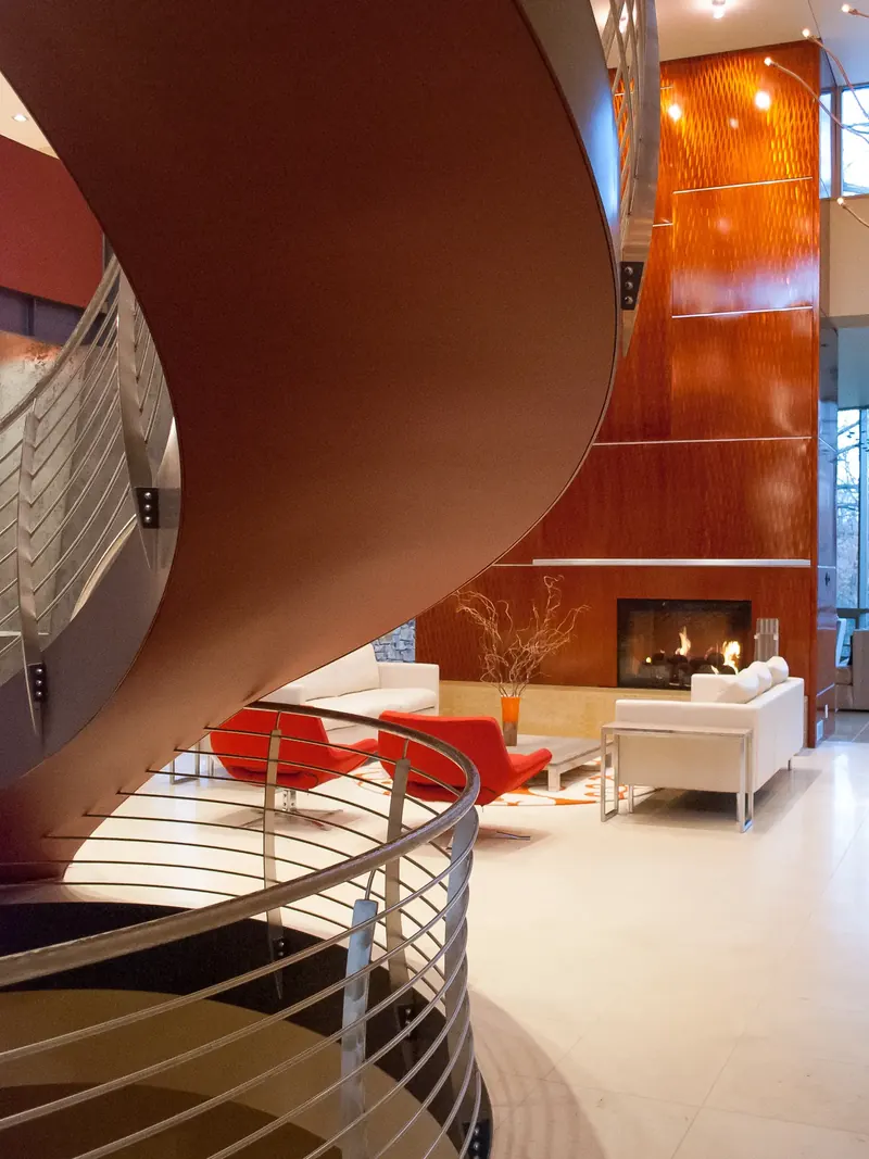 Underside of the red spiral stair framing the living room, white sofas, red chairs and a tall fireplace wall