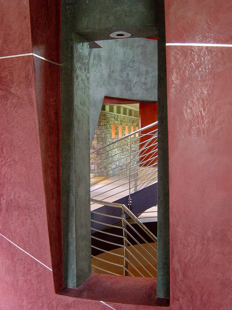 Narrow slot cut through a red plaster privacy wall, revealing a glimpse of the steel stair railing and stone wall beyond