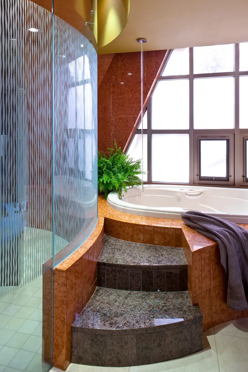 Stone steps rising to a tub clad in red stone tile, curved etched-glass shower wall at left