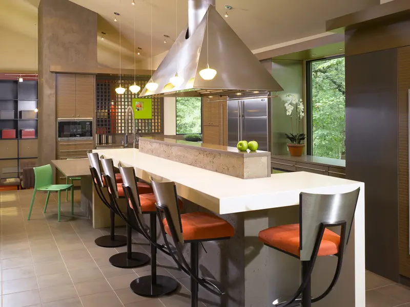 Long island with orange-seated stools beneath a sculptural stainless steel range hood