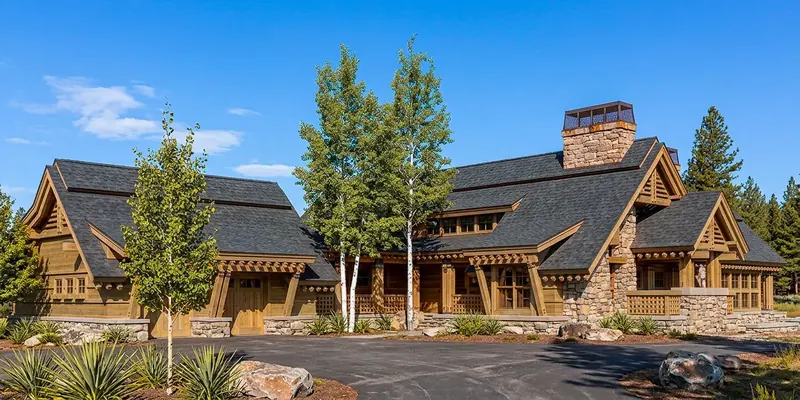 Wide front elevation with timber porches, stone masonry, white-barked trees and a curved driveway