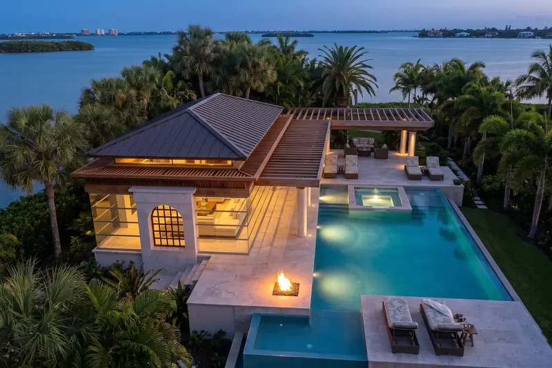 Aerial view at dusk of the Modern Classical Pavilion, its lit pool and pergola terrace set among palms beside open water