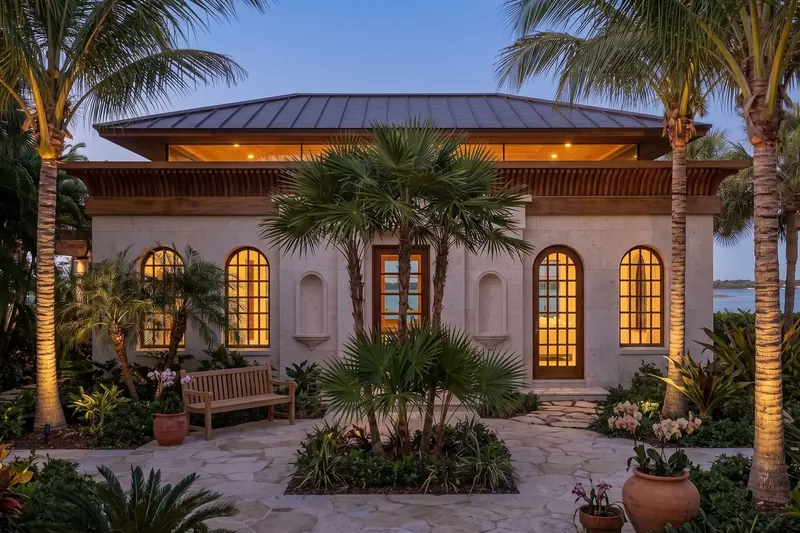 Stone guest house facade with arched wood windows, niches and a central door behind a palm garden