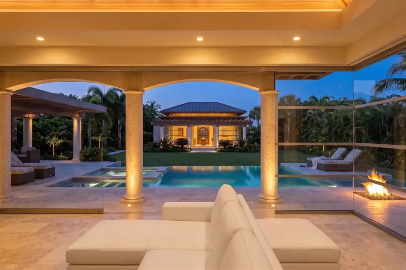 From a white sofa inside, the view through stone arches across the pool to a lit building beyond the lawn