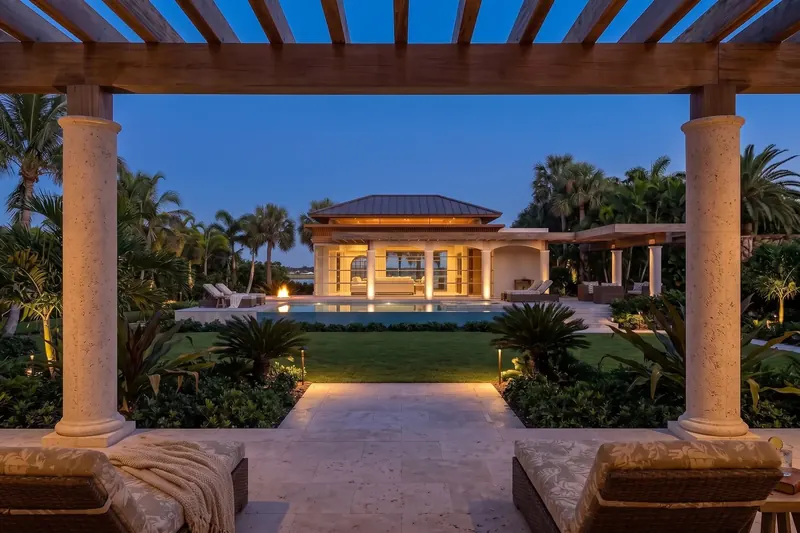 View between two stone columns under a wood pergola, across the lawn to the lit pavilion and pool