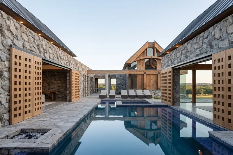Central courtyard pool framed by stone pavilions with open gridded barn doors, loungers and a timber gable beyond