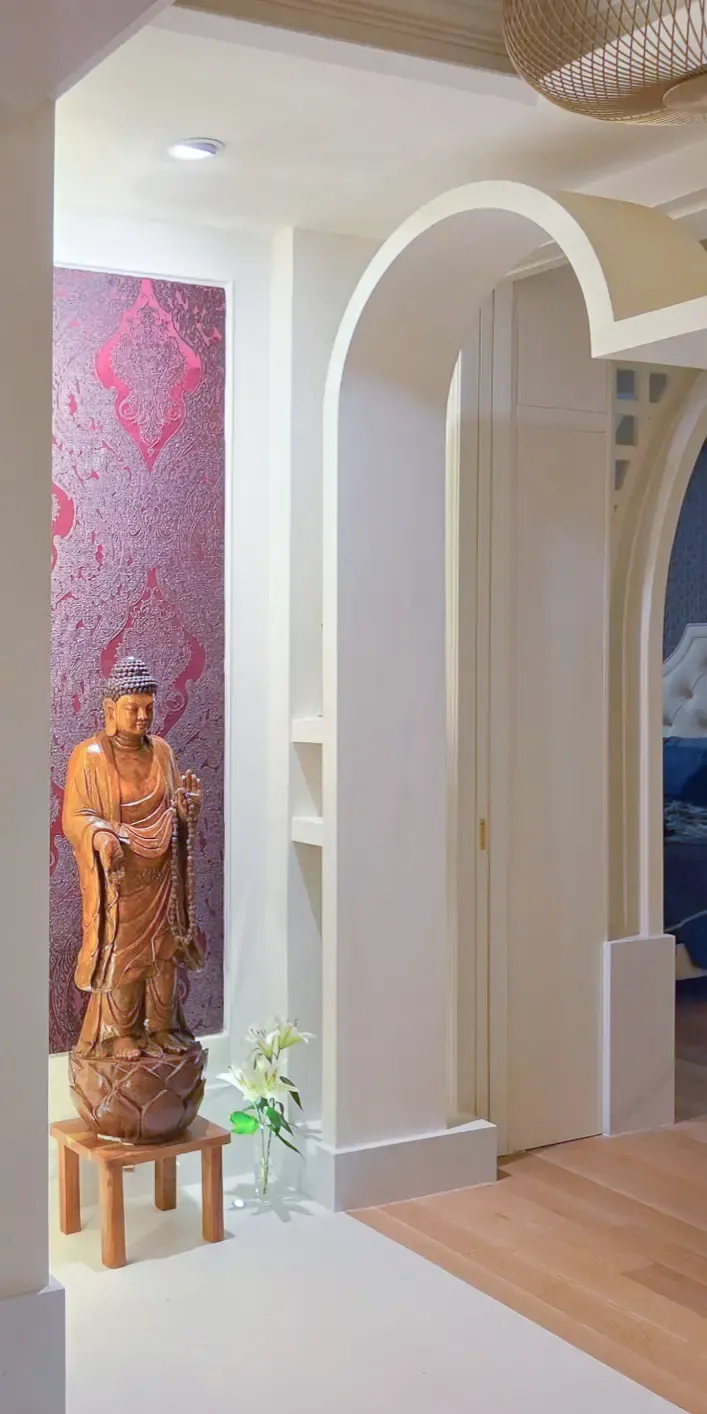 Softly lit wooden Buddha statue and white lilies in a niche with damask wallcovering, beside a white arched passage