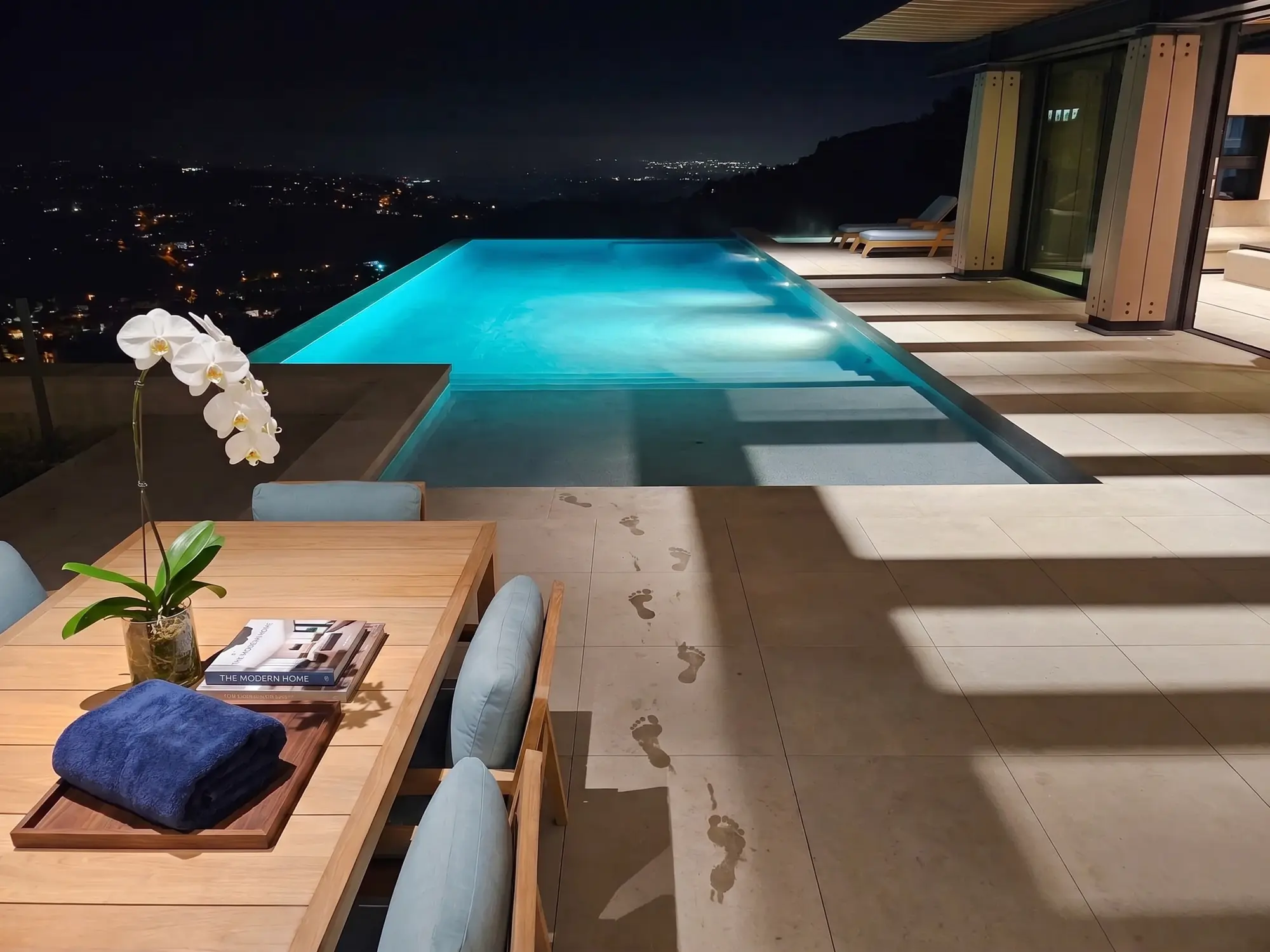 Wet footprints lead from the outdoor table to the glowing infinity pool above the city lights