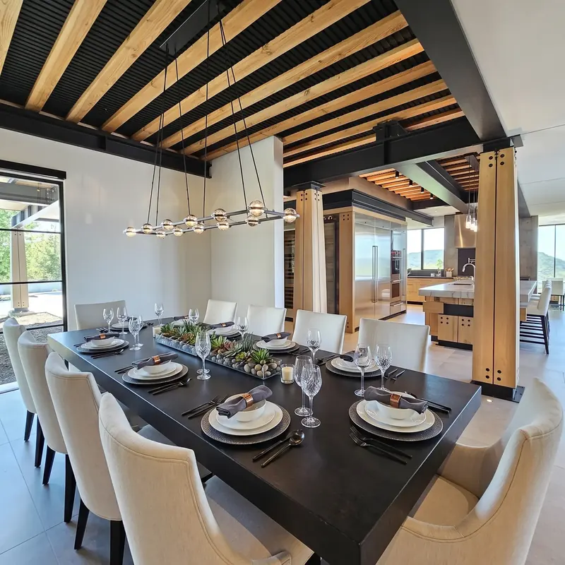 Long dining table set for guests beneath exposed timber beams, open to the kitchen beyond