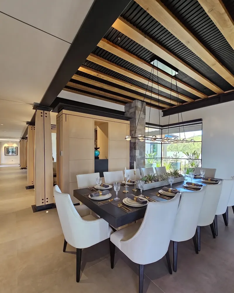 Dining room under timber beams and black slats beside a gallery of bolted columns and a stone pier