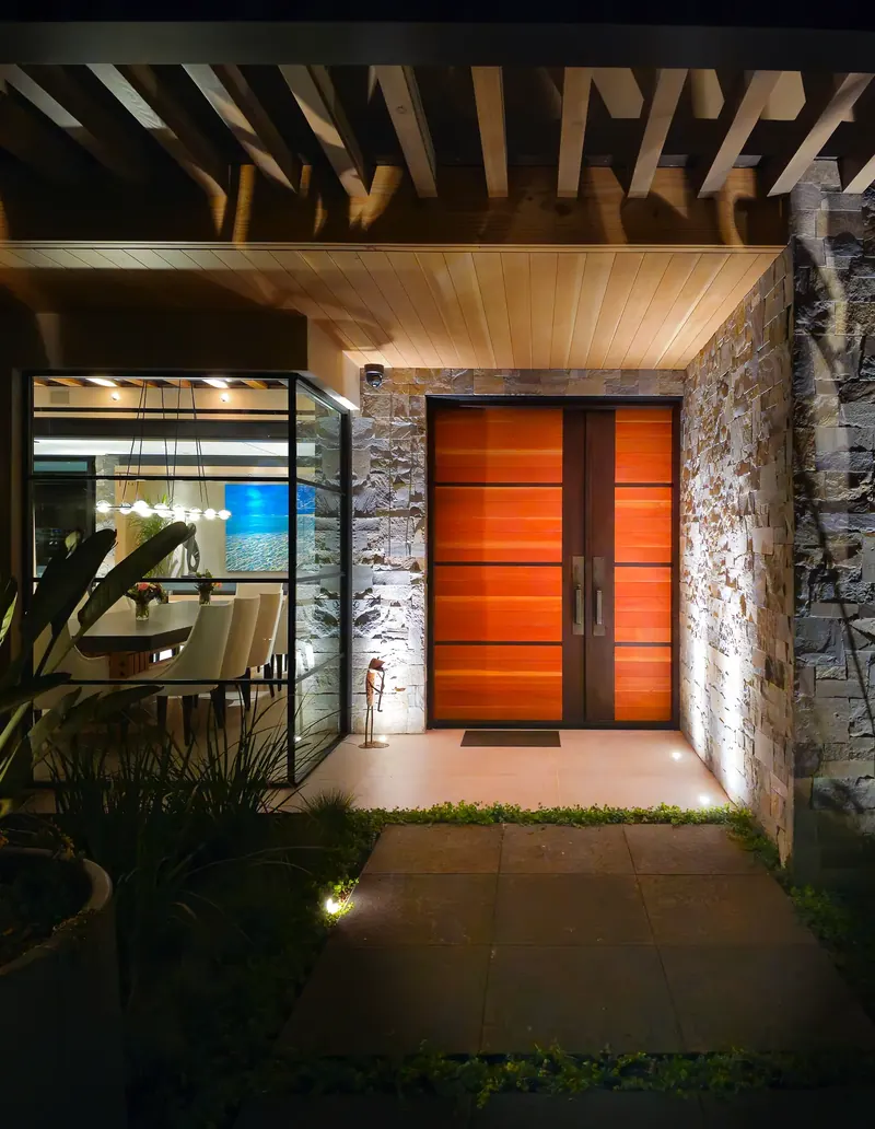 Front entry at night: a wood double door with horizontal bands, set in stone beneath a timber trellis