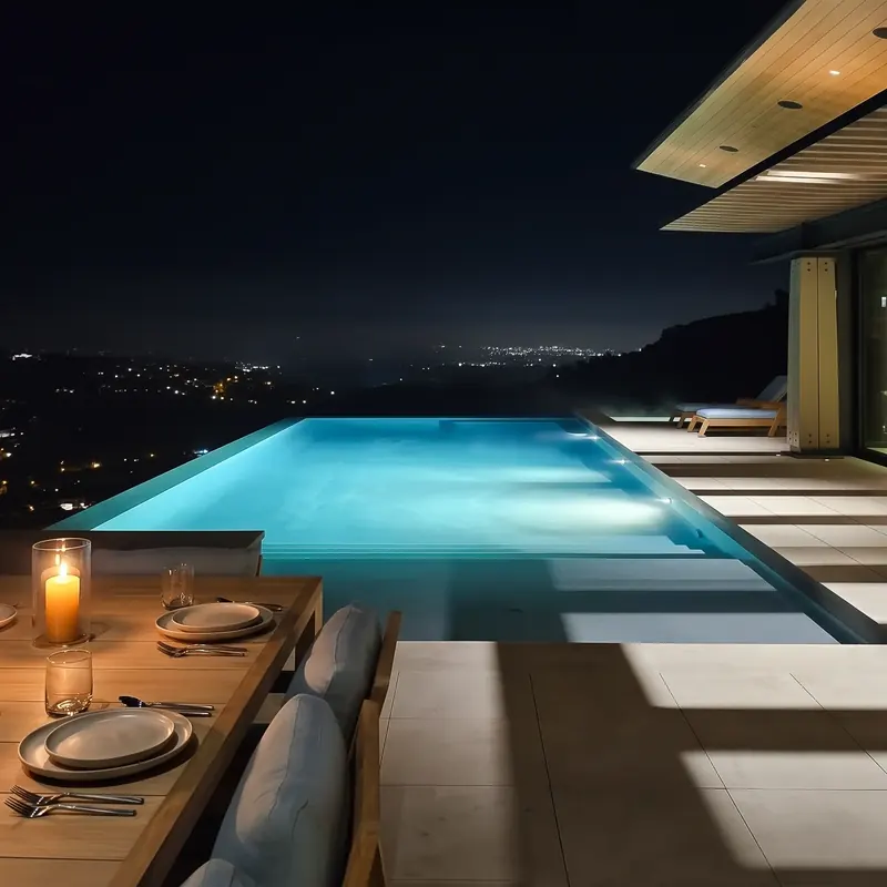 Lit infinity-edge pool at night beside a candlelit outdoor table, city lights below
