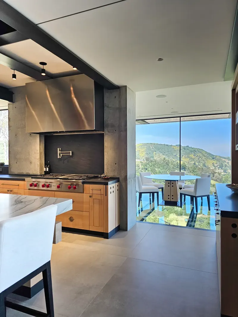 Cooktop recessed in an exposed concrete wall, the glass dining room and green hillside to the right