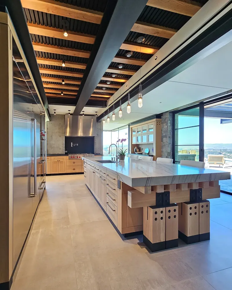 Long kitchen island on timber trestle legs beneath a ceiling of exposed beams and black slats