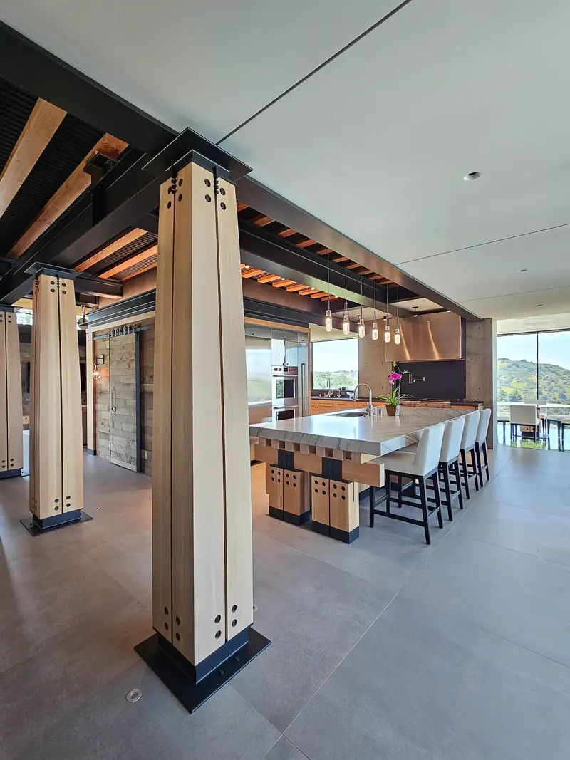 Bolted timber columns frame the kitchen island, with the pantry, cooking wall and glass dining room beyond
