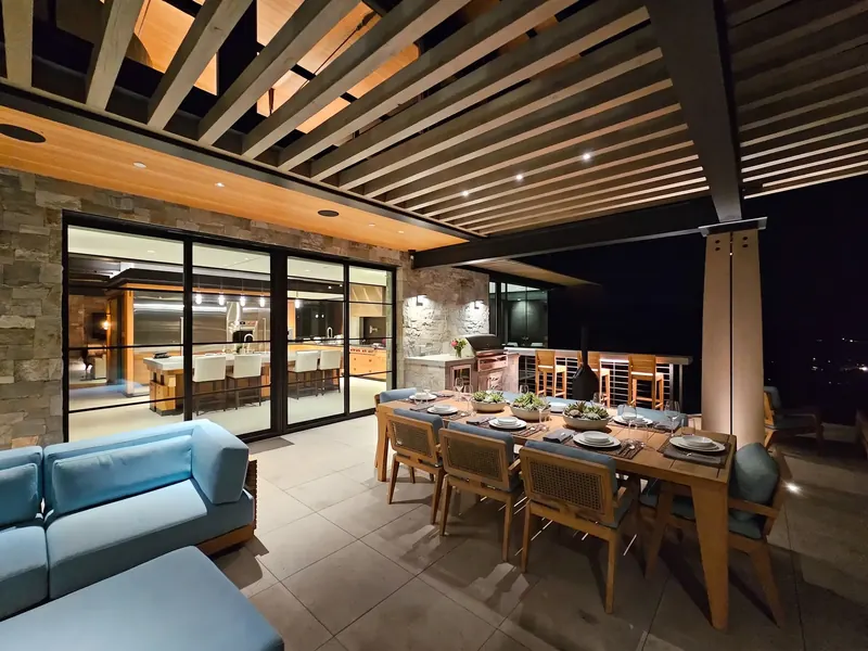 Outdoor dining and lounge at night under the lit trellis, the kitchen visible through glass doors