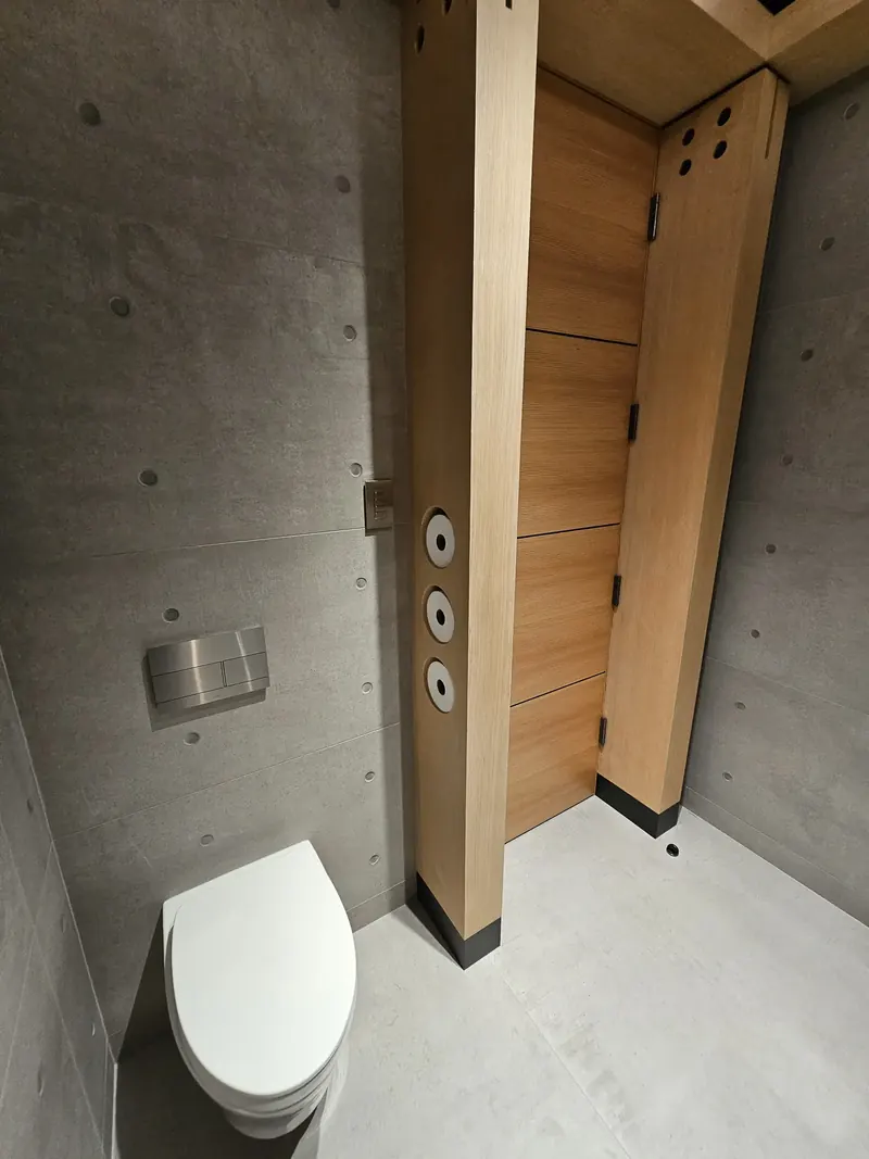Powder room of concrete panels with a wall-hung toilet and a wood column holding paper rolls