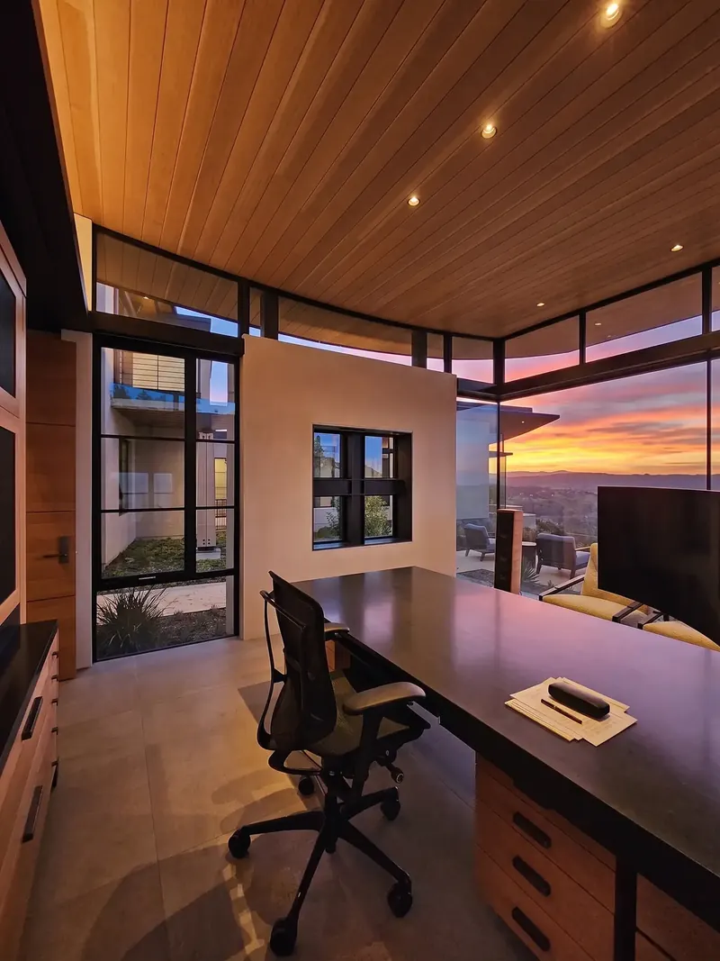 The study at sunset under a curved wood ceiling, the sky glowing through corner glass beyond the desk