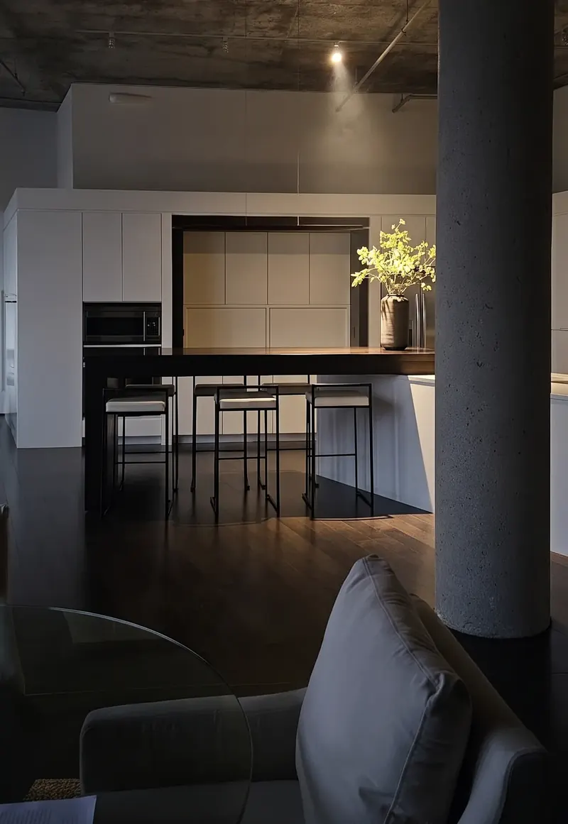 Dimly lit view past a sofa to the dark bar counter and stools, one spotlight on a vase of greenery, concrete column at right