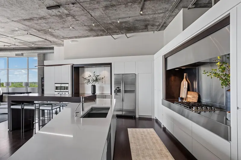 White kitchen with long island and sink facing a cooking alcove lined in stainless steel under a concrete ceiling