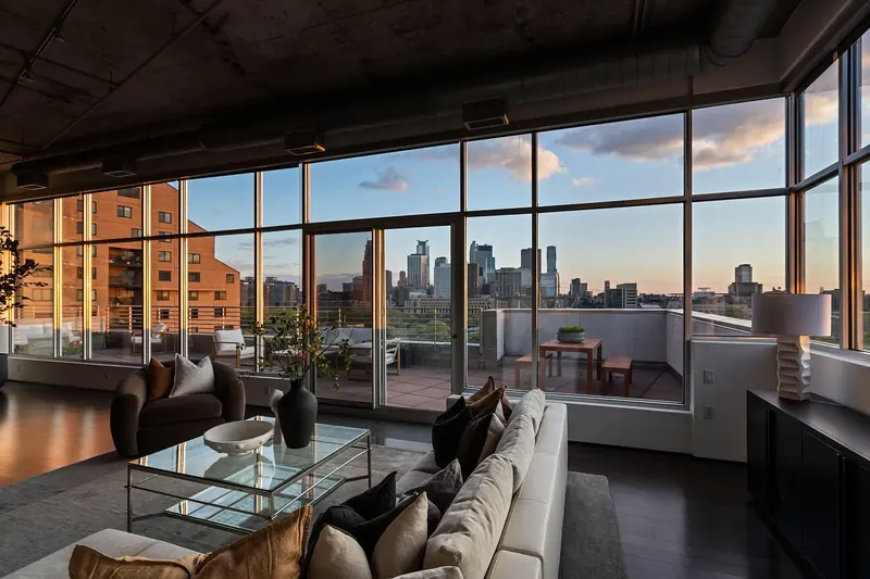Sofa and glass coffee table before corner windows and a terrace, with the downtown skyline in late sun