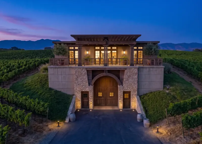 The Sonoma Vineyard Estate at dusk, lit windows above arched wooden winery doors set into a vine-covered hillside