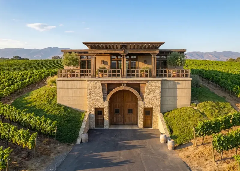 Front elevation in daylight: arched double doors in stone below a balcony with potted olive trees, flanked by earthen berms