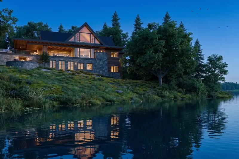 Stone & Timber Lake Home at dusk, its glass gable and lit windows reflected in the lake below a grassy slope