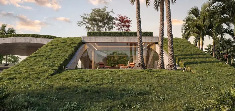 Glass-walled living space set into a grass-covered berm beneath a concrete lintel, with palms in front at sunset