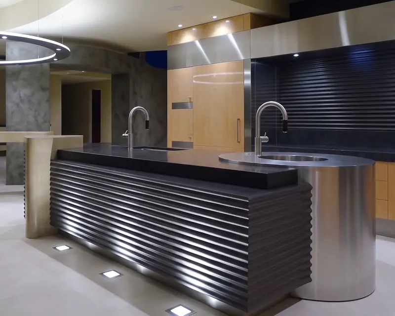 Center island wrapped in wavy black material, ending in a stainless steel drum sink, with floor lights along its base