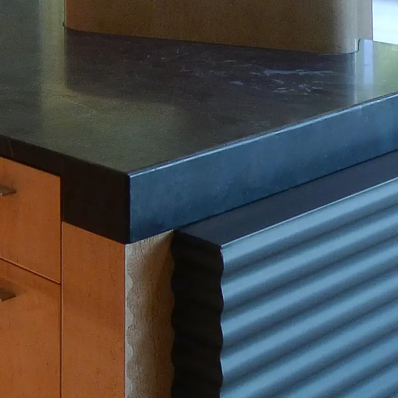 Corner detail where a thick dark countertop meets maple cabinetry and the wavy black island cladding
