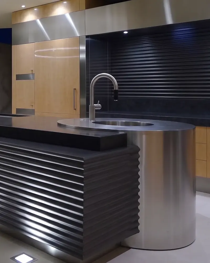 Cylindrical stainless steel sink drum meeting the corrugated black island, with a ribbed backsplash behind
