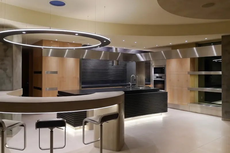 The Lair kitchen at night beneath a suspended ring light, with a curved concrete bar, stools and a corrugated black island