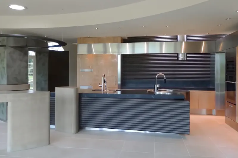 Daylight view of the kitchen under a curved ceiling, with concrete bar forms, birdseye maple panels and the black island