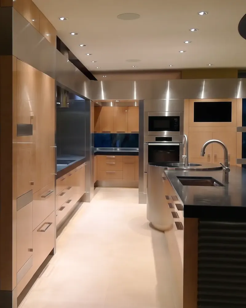 View past the island's stainless drum sink toward maple cabinetry, built-in ovens and a continuous stainless band