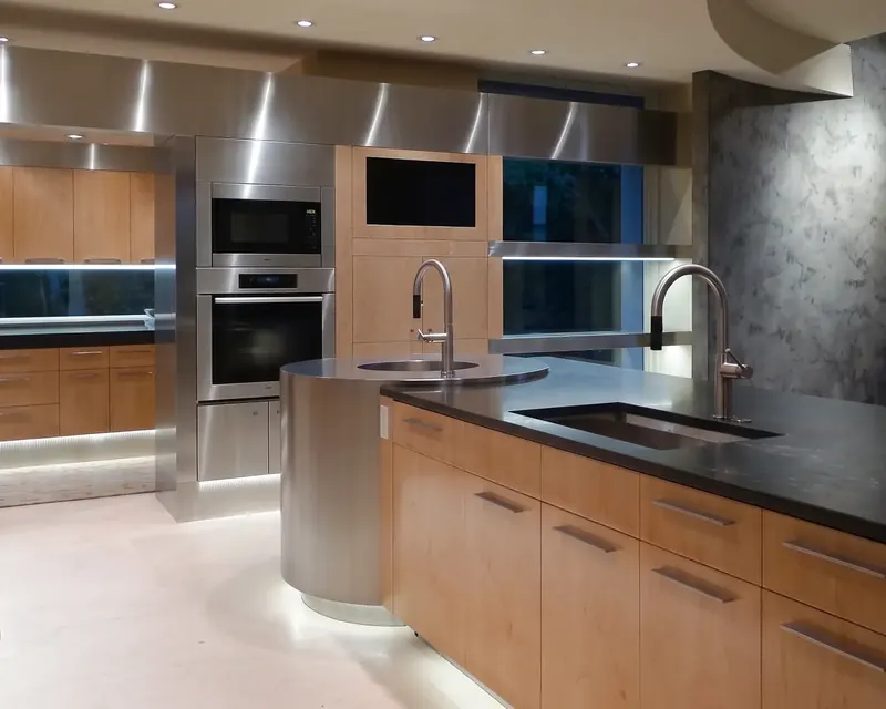 Island with maple drawers, a dark countertop and two sinks, the stainless drum sink glowing above an illuminated base