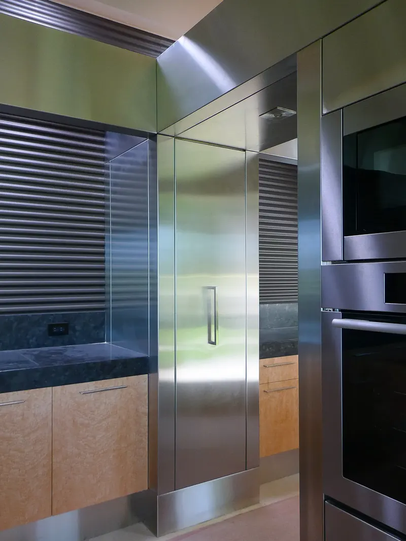 Stainless steel clad tall cabinet between a corrugated black backsplash and built-in wall ovens
