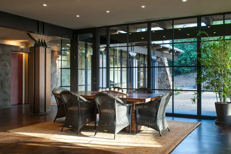 Dining room with wicker chairs and a timber table before a wall of steel-framed glass