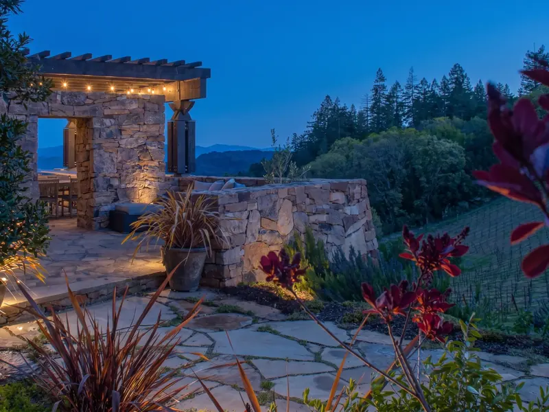 Stone courtyard and pergola at dusk, with string lights and red flowers