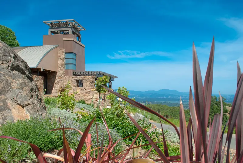 Stucco tower and curved metal roof rising behind a boulder and planted slope, with the valley beyond