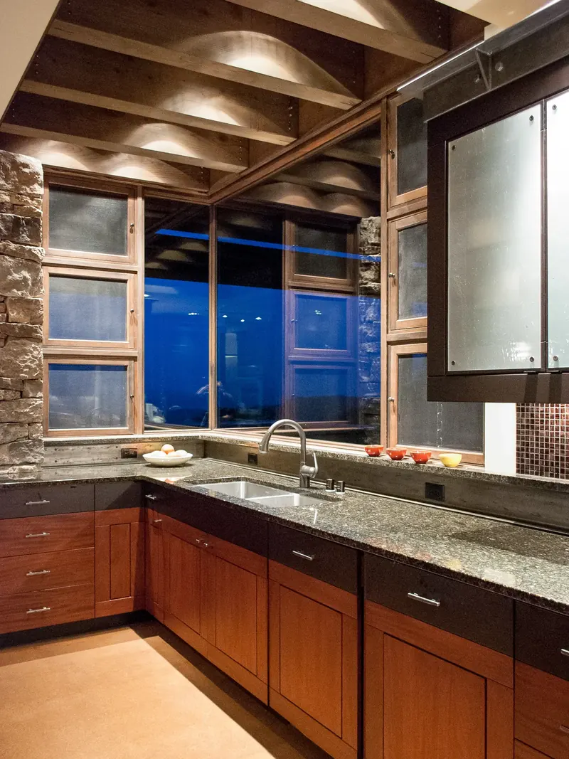 Kitchen sink beside a stacked stone wall, with a timber ceiling and windows to the evening sky