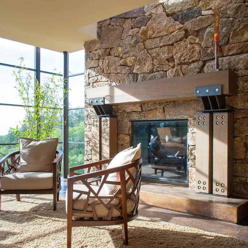 Stone fireplace with heavy timber and steel mantel, flanked by armchairs and tall windows