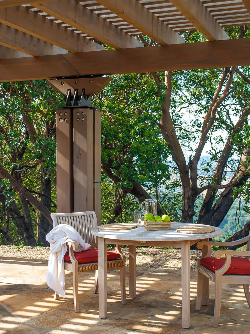Round outdoor table with red-cushioned chairs beneath a timber pergola among trees