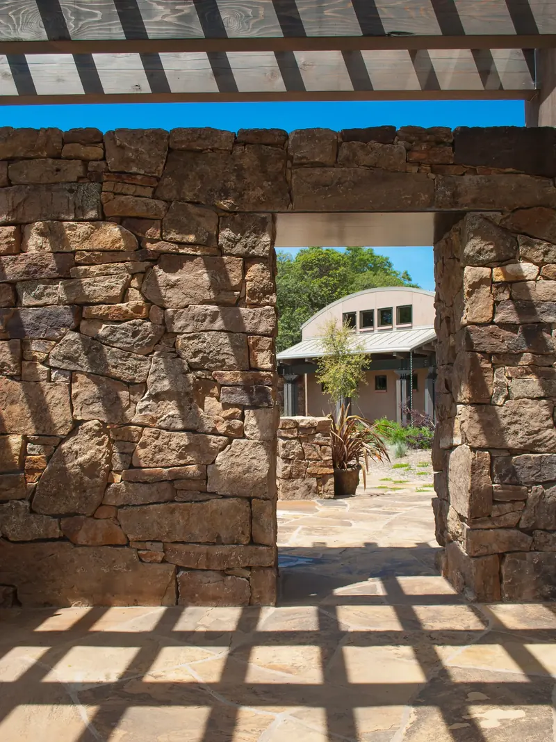 Stone wall opening framing a courtyard, with pergola shadows cast across the flagstone
