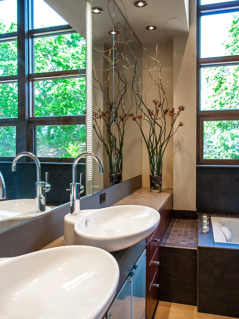 Twin white basins on a dark counter before tall windows to the trees
