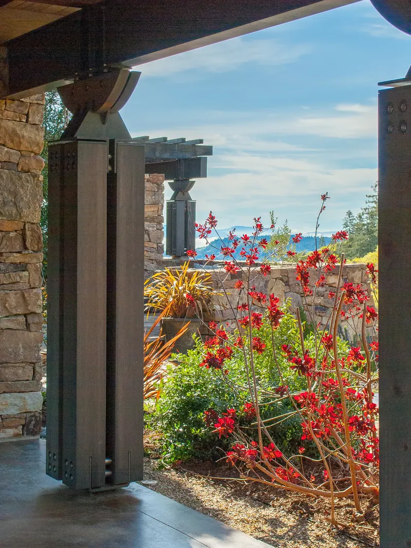 Covered walkway with steel-clad posts opening to red flowering plants and distant hills
